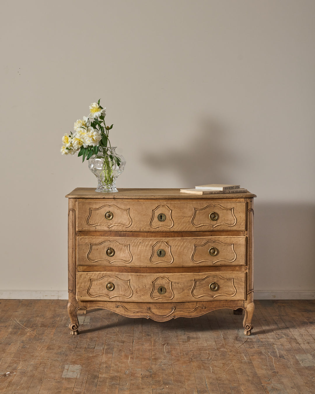 Light wood commode with three carved drawers, topped by a vase of white flowers and books.
