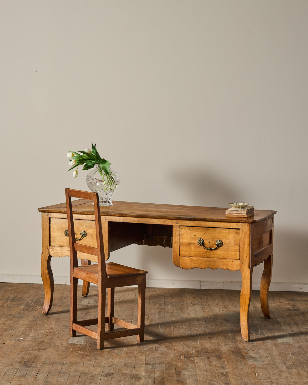 A wooden desk with two drawers, a simple wooden chair, a vase of white tulips, and a small box on a wooden floor.