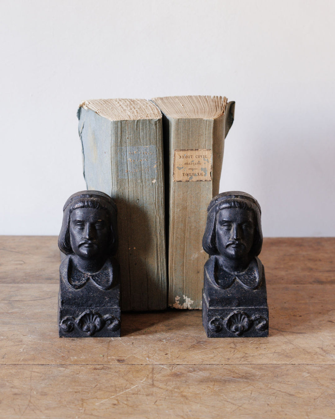 Two black bookends shaped like men's busts, flanking two old light blue books on a wooden table.