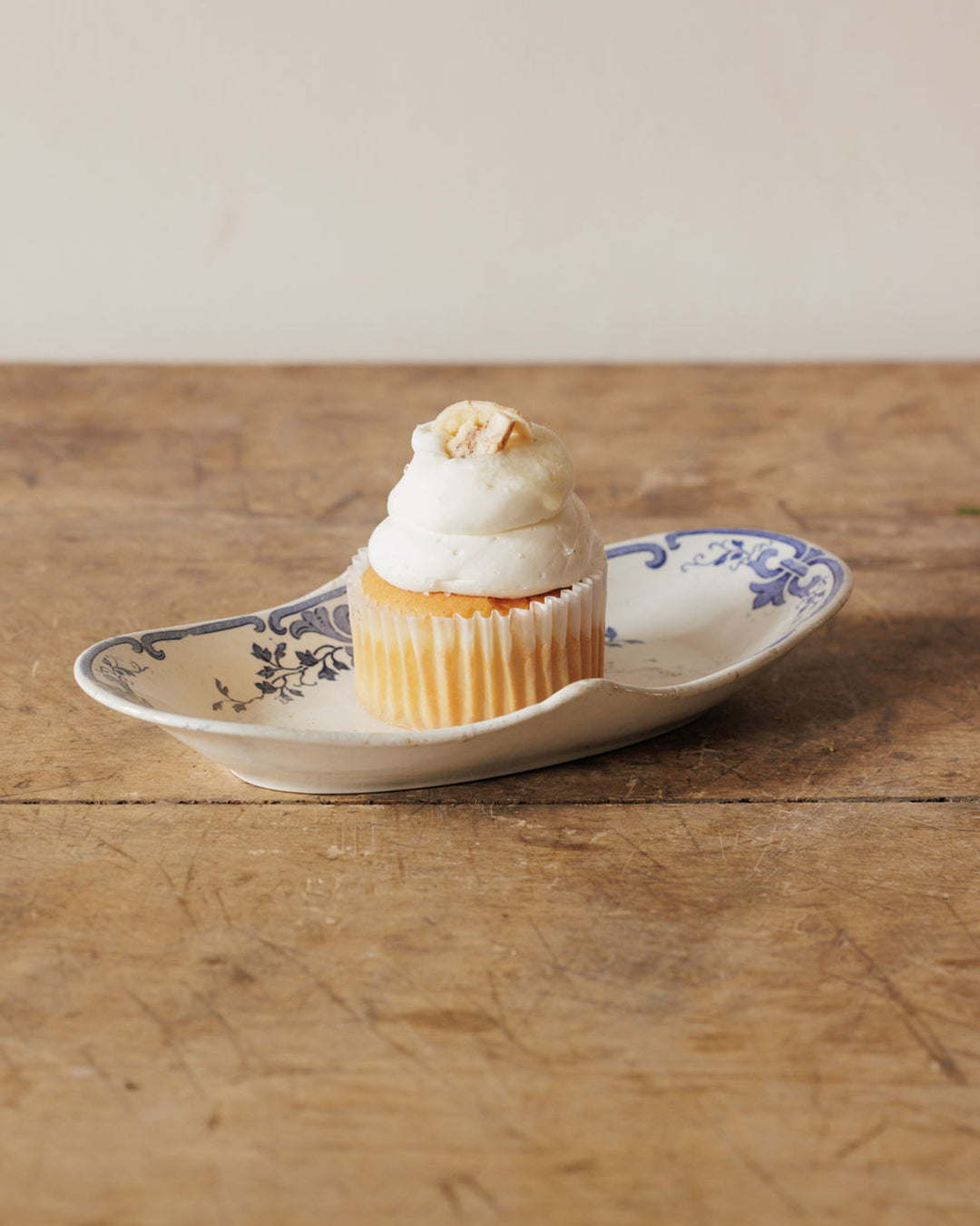 A cupcake with white frosting and a nut topping on a patterned dish on a wooden surface.