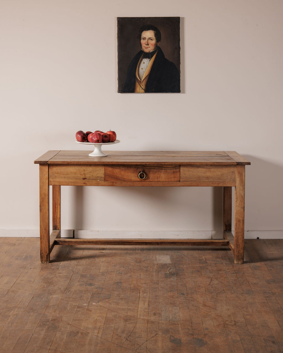 A rustic wooden console table with red apples on a stand, beneath a framed portrait painting of a man.