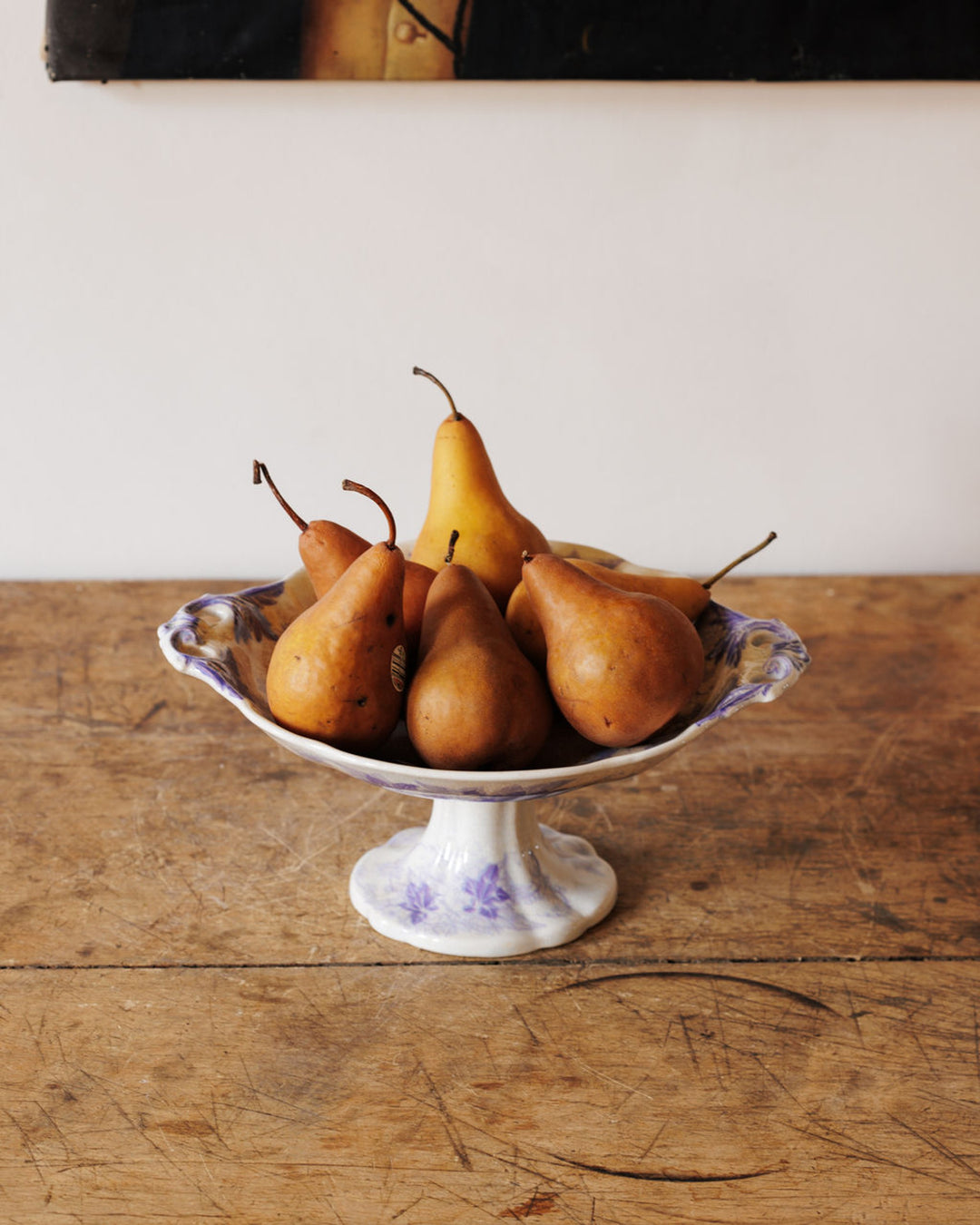 A white pedestal bowl with purple floral designs holds an assortment of ripe reddish-brown and yellow pears on a rustic wooden table.