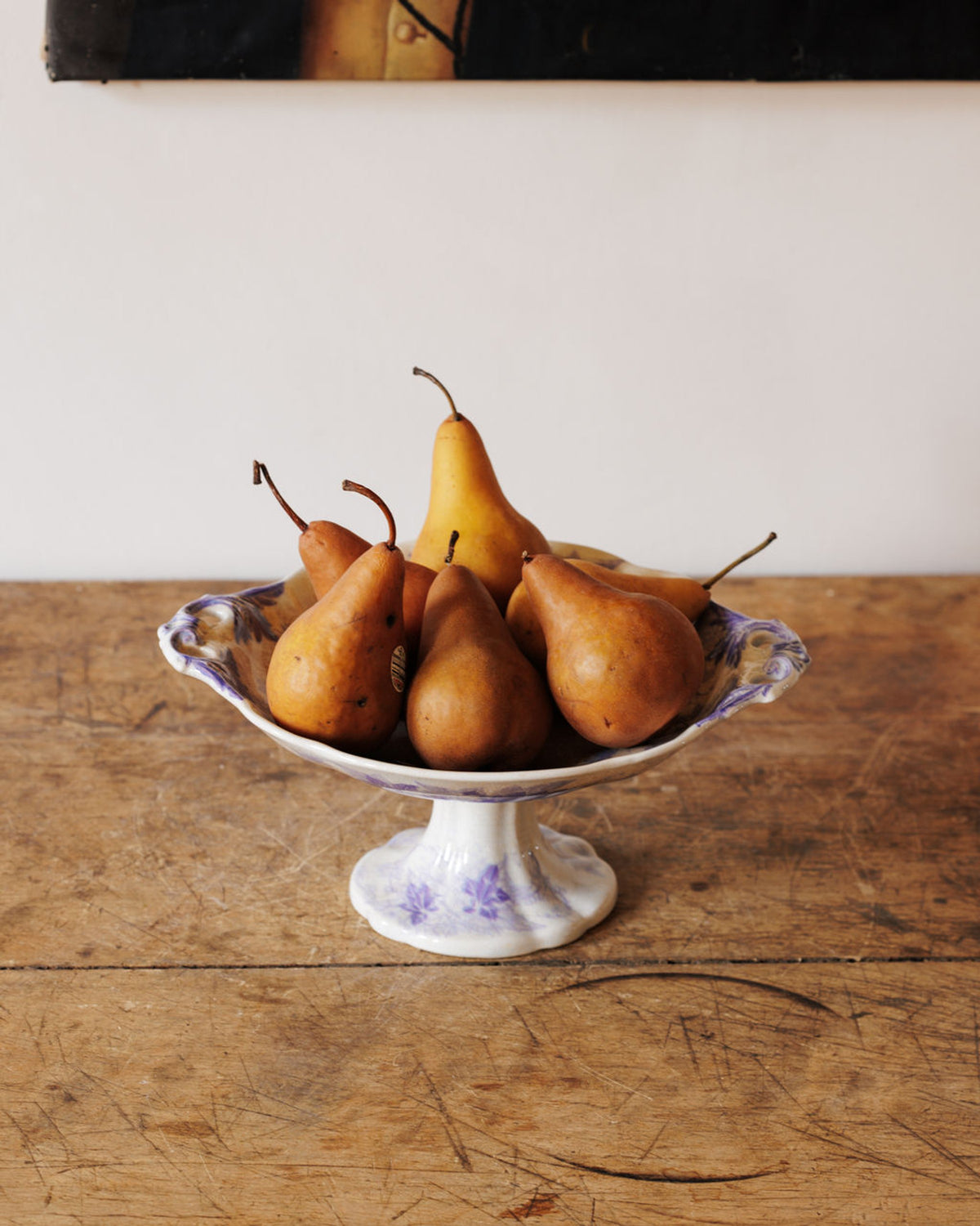 A white pedestal bowl with purple floral designs holds an assortment of ripe reddish-brown and yellow pears on a rustic wooden table.