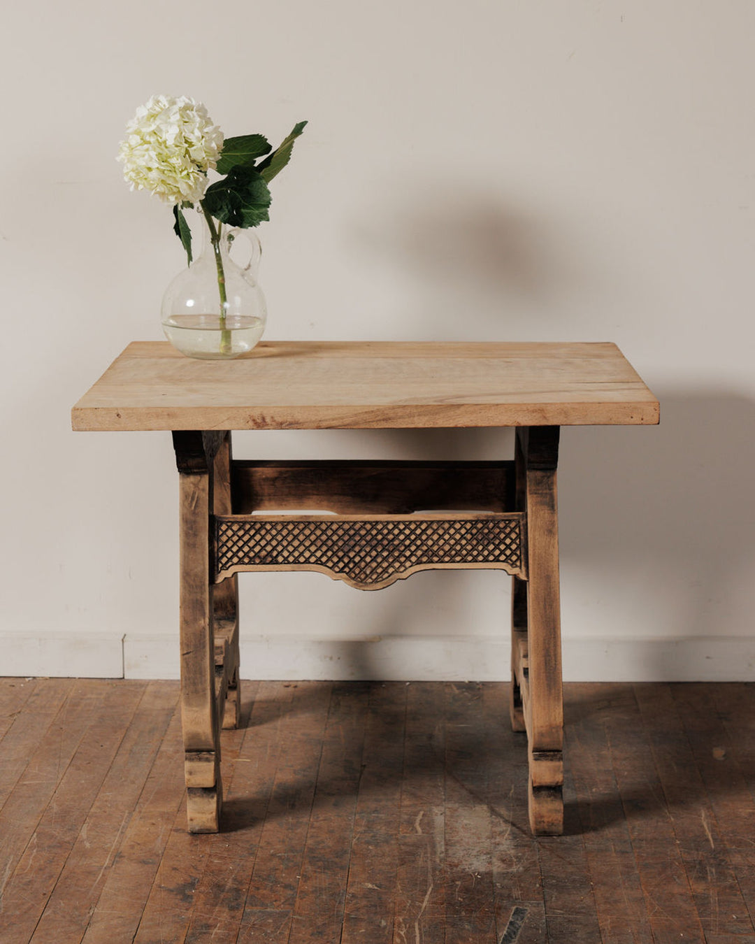 A rustic wooden side table with carved details, holding a clear vase with a white hydrangea.