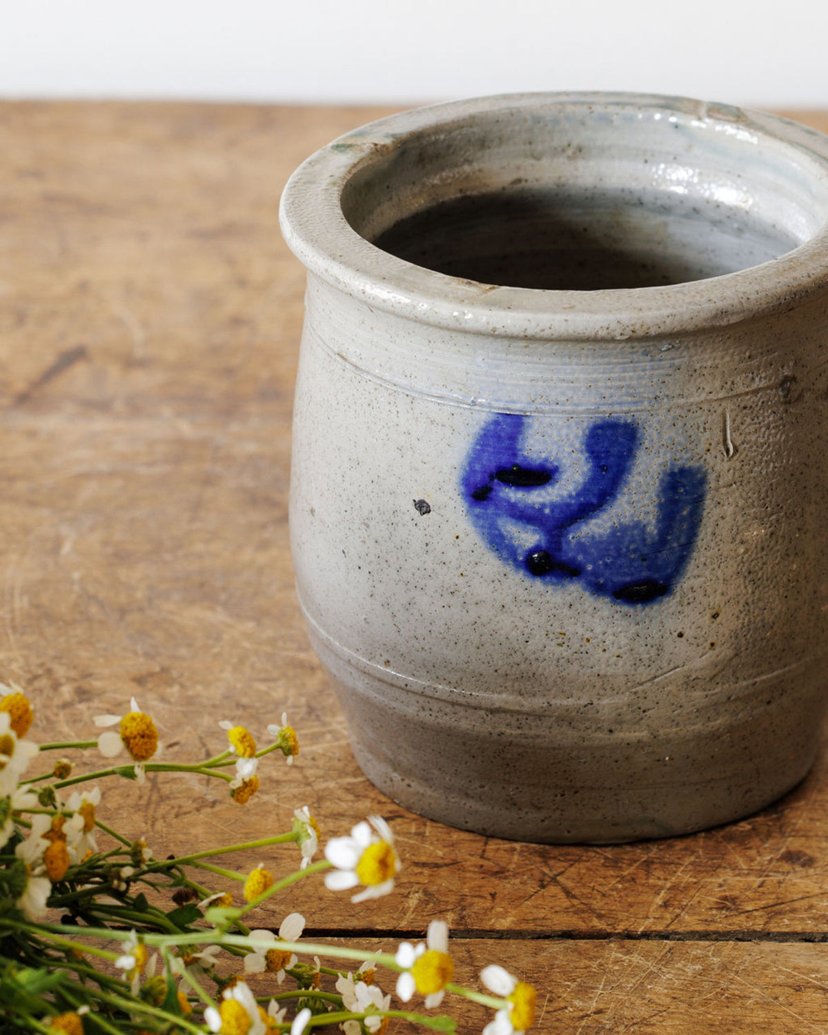 Speckled grey stoneware crock with blue swirl design on a wooden surface, beside white and yellow wildflowers.