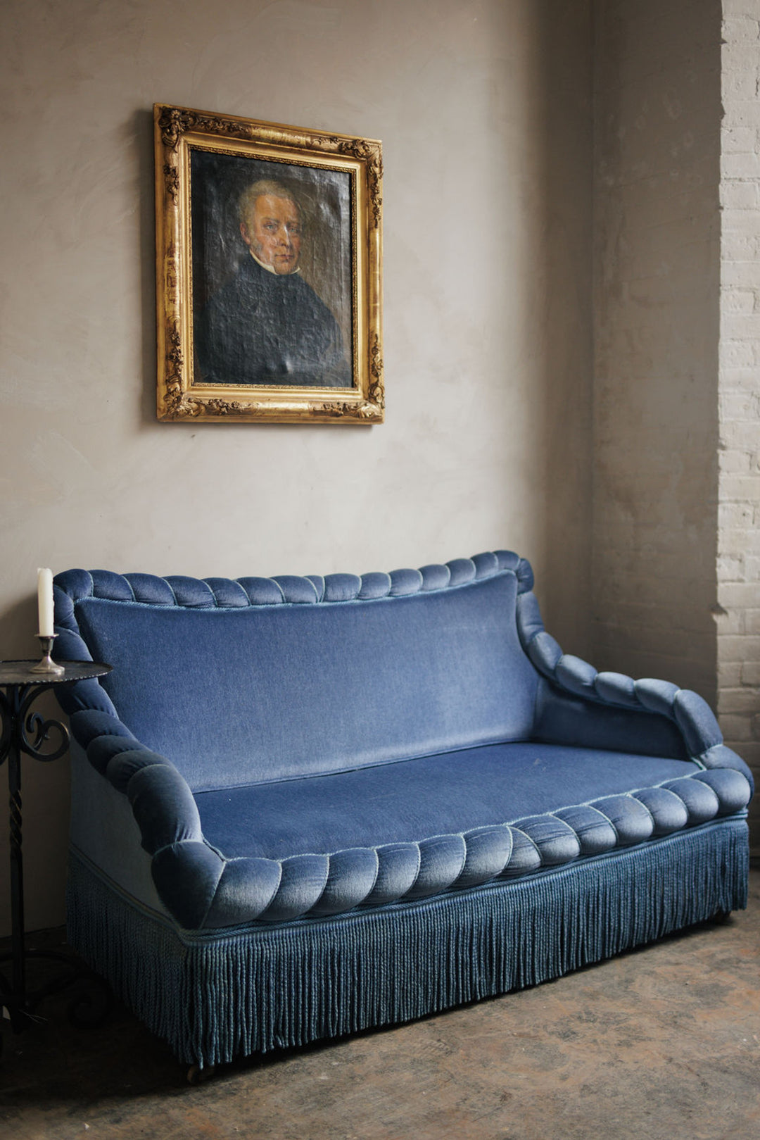 A blue velvet sofa with braided trim and fringe skirt sits below an antique portrait in a gilded frame.