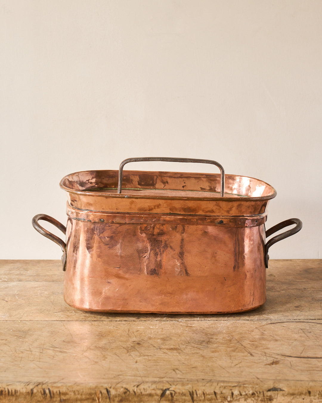 An oval copper fish kettle with a removable internal rack sits on a wooden table.