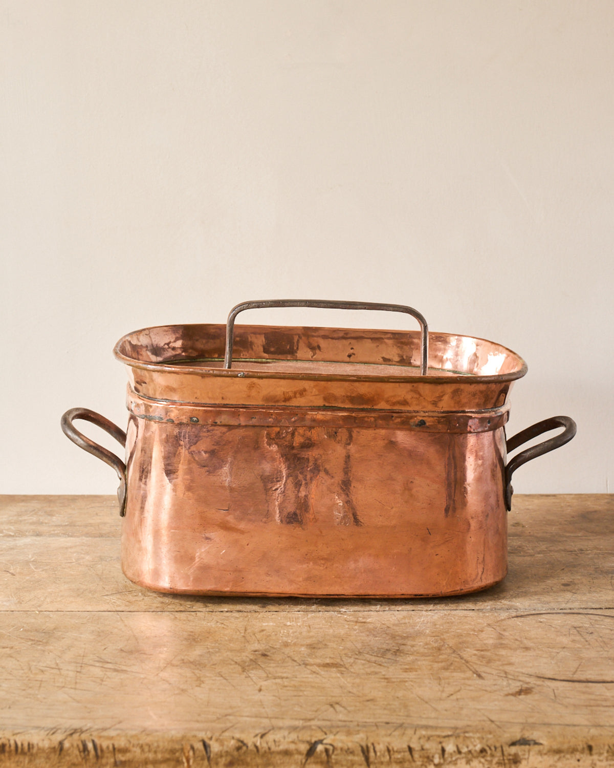 An oval copper fish kettle with a removable internal rack sits on a wooden table.