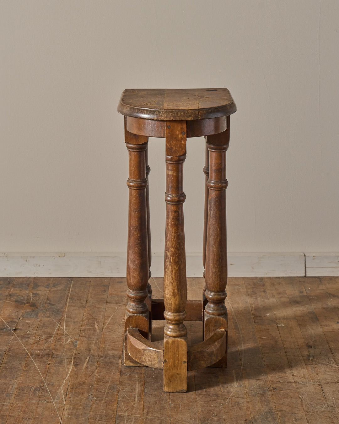A tall, D-shaped wooden stool with turned legs stands on a worn wooden floor.