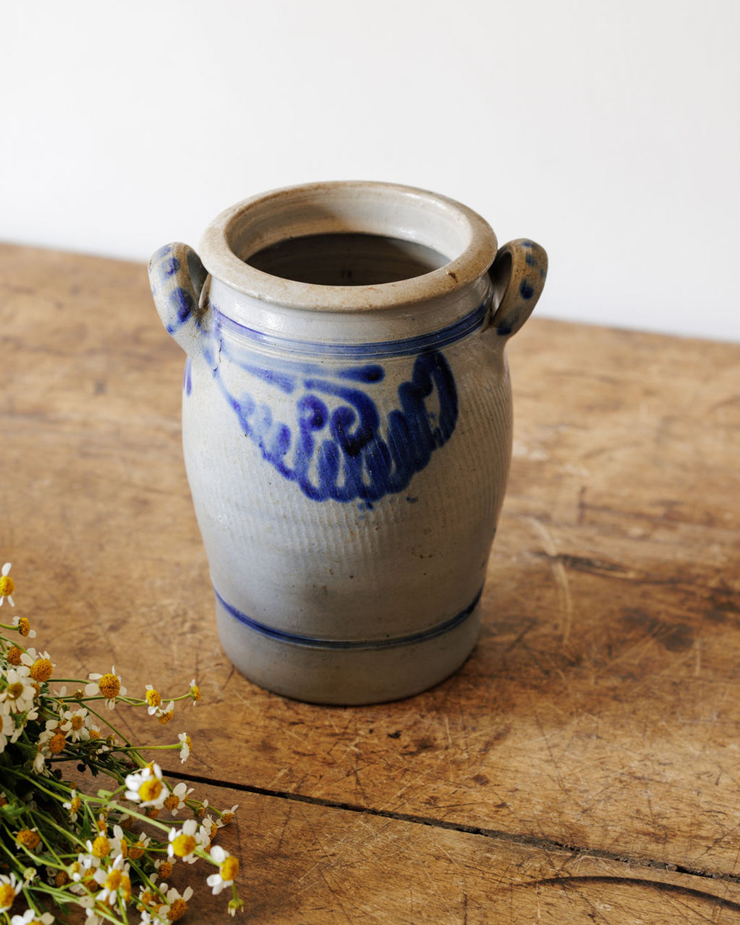 A gray stoneware crock with blue floral decoration and two handles, sitting on a wooden table with small white flowers in the foreground.