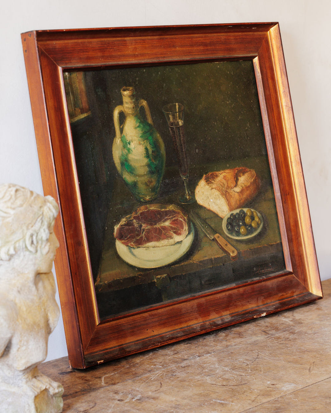 A framed still life painting depicting a green jug, wine, bread, meat, and olives, next to a white bust.